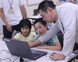子供達の前に置かれたノートパソコンを操作している大人と、それを楽しそうに見ている二人の子どもたちの写真