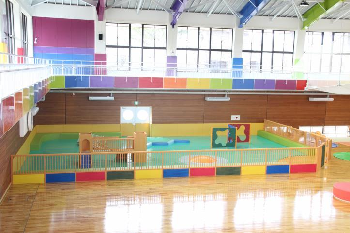 広い施設の一画に木製のフェンスで囲まれ、中に遊具やマットが設置されてある写真