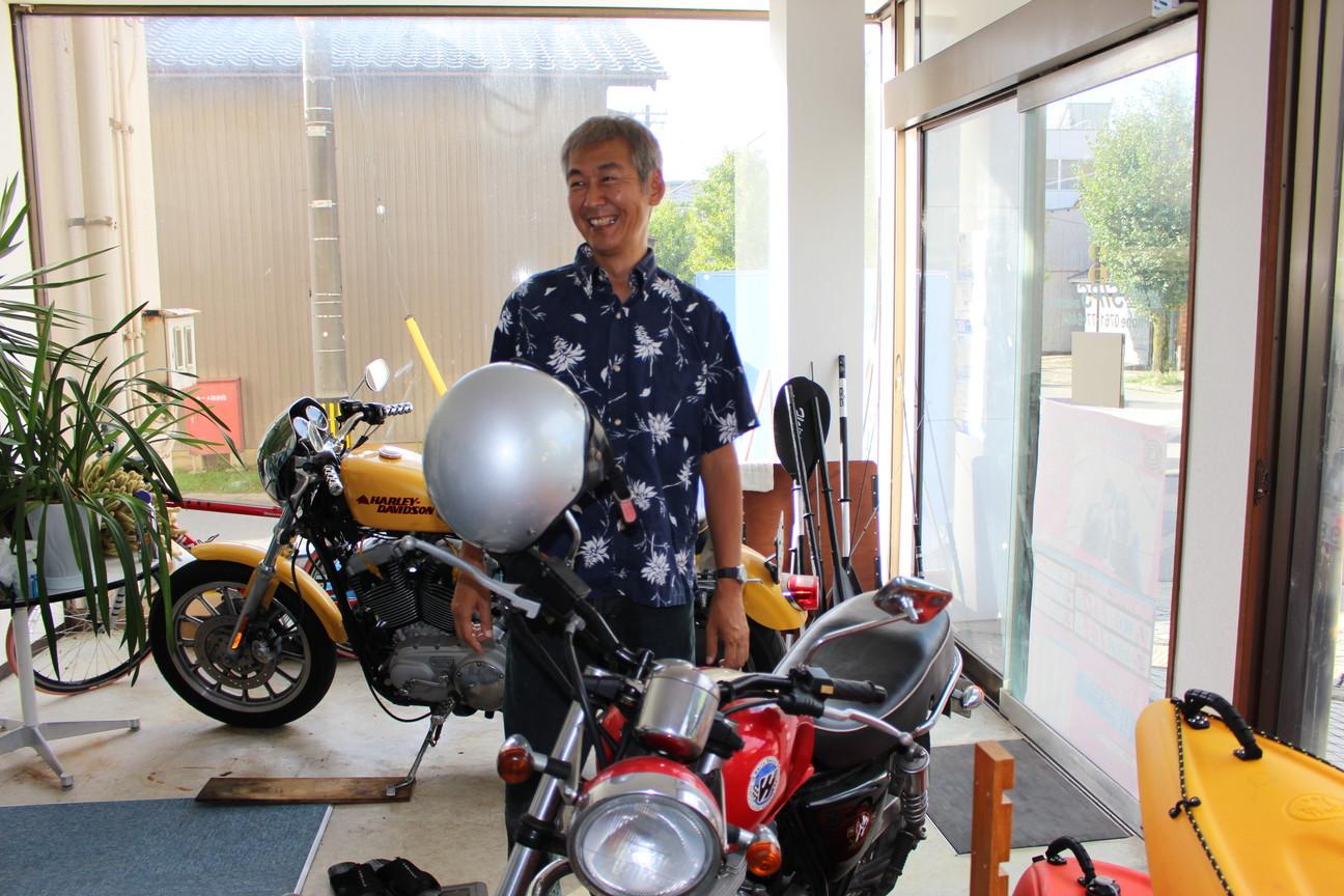 バイクが2台とカヌーが置かれた室内で笑顔で立っている久保田雅裕さんの写真