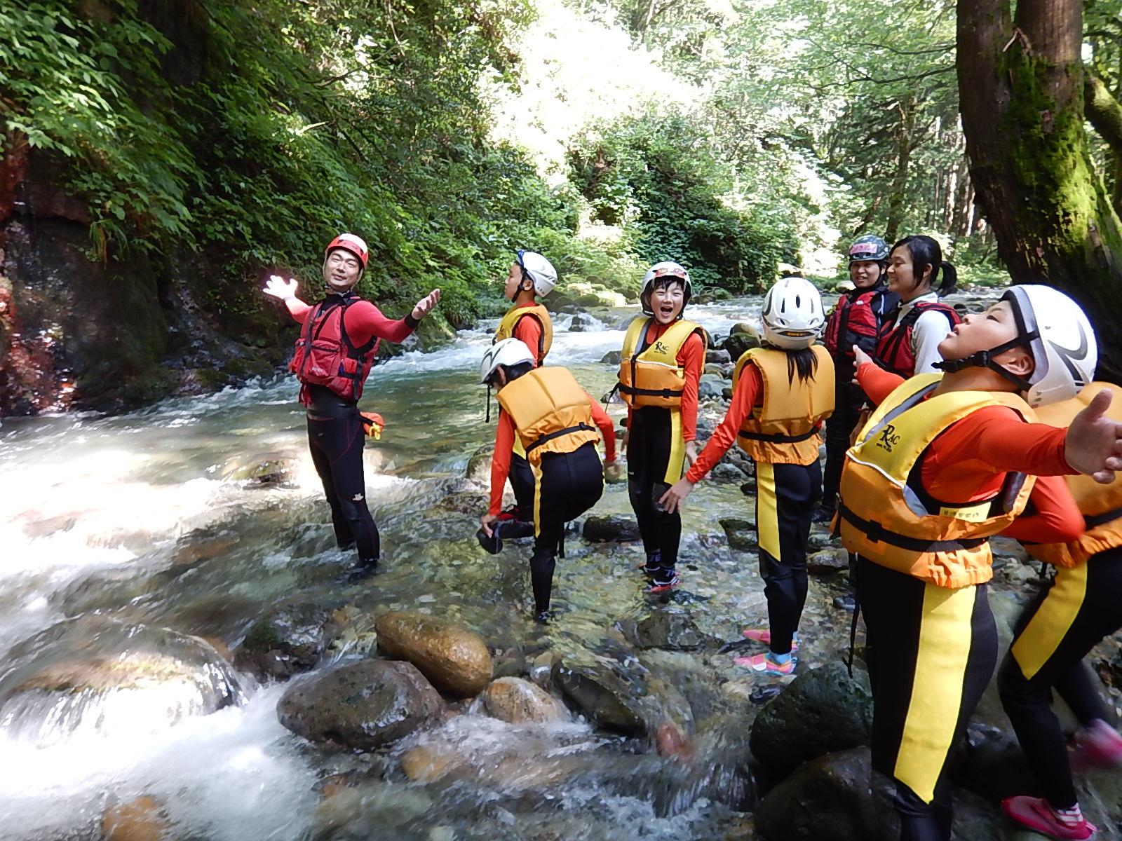 森の中の小川で救命胴衣を着けて両手を広げている増本さんと子供たちの写真