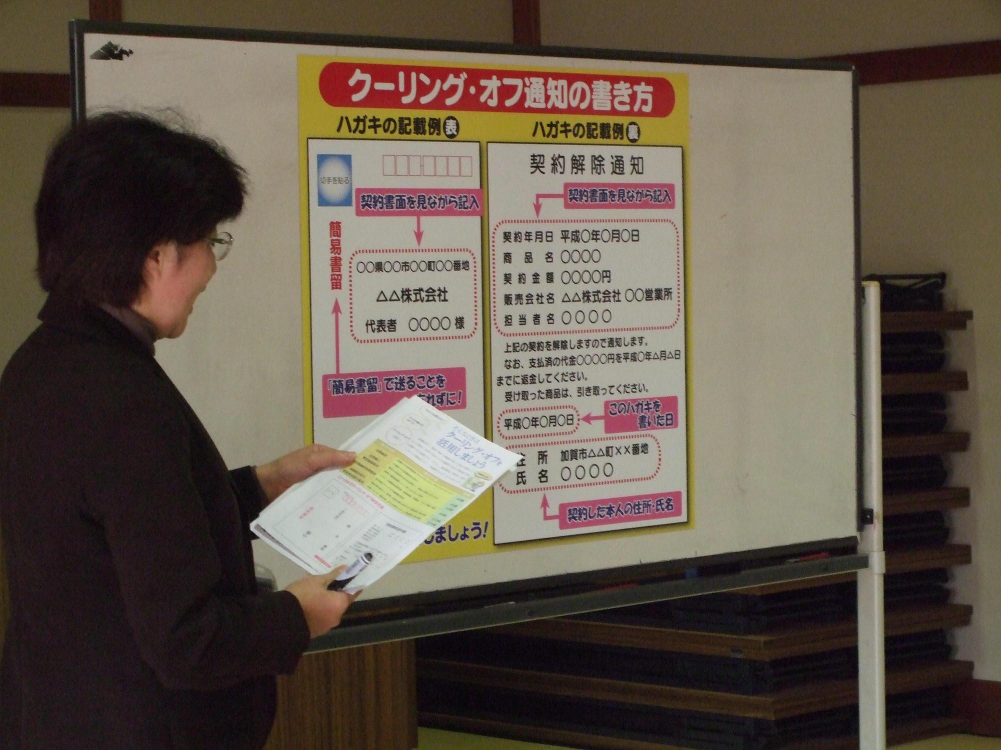 ホワイトボードに貼られたクーリング・オフ通知の書き方の紙の横で説明をしている女性の写真