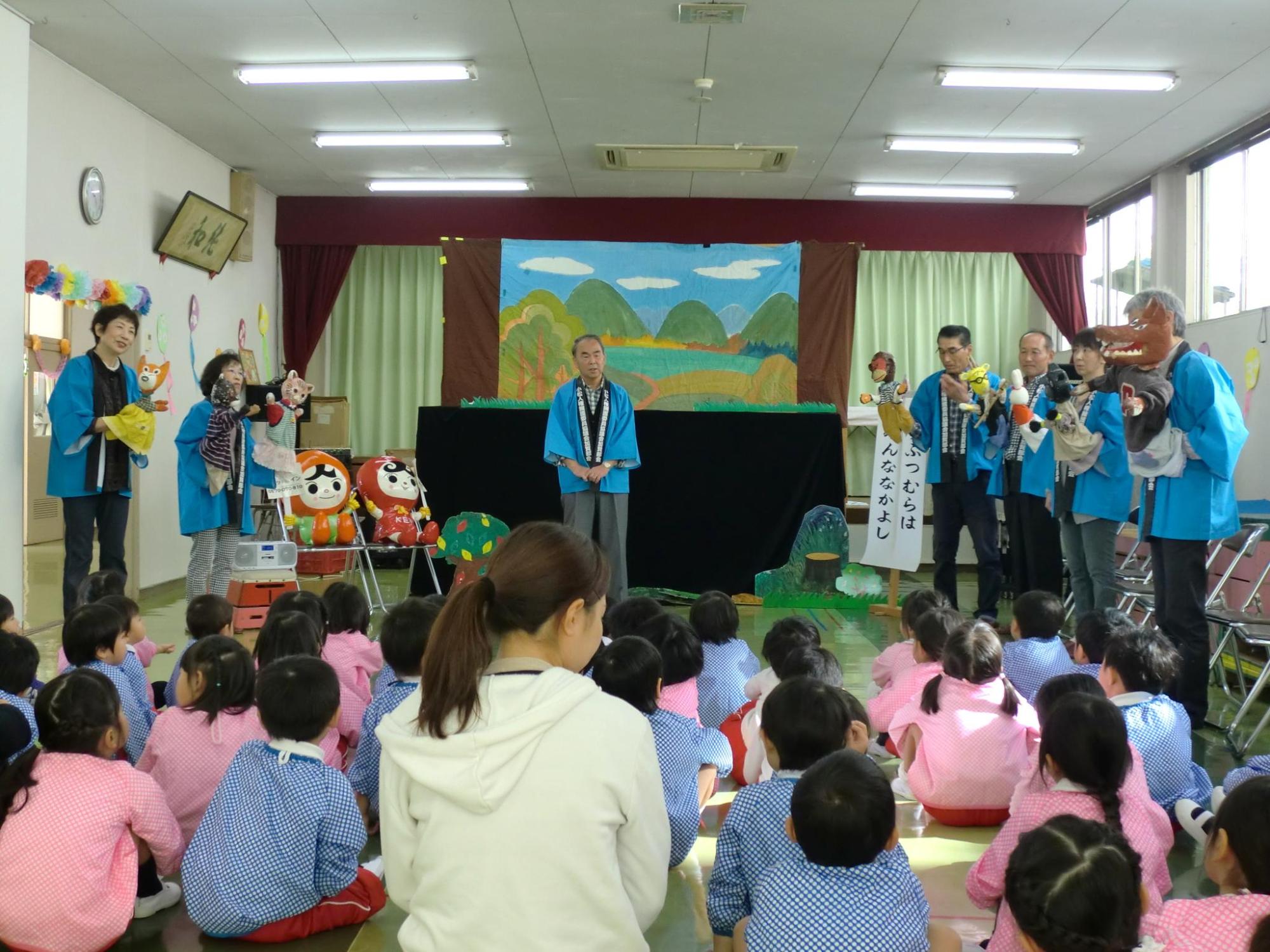 青い法被を着た人形劇のスタッフさんたちが人形を持って子供たちの前に並んでいる写真