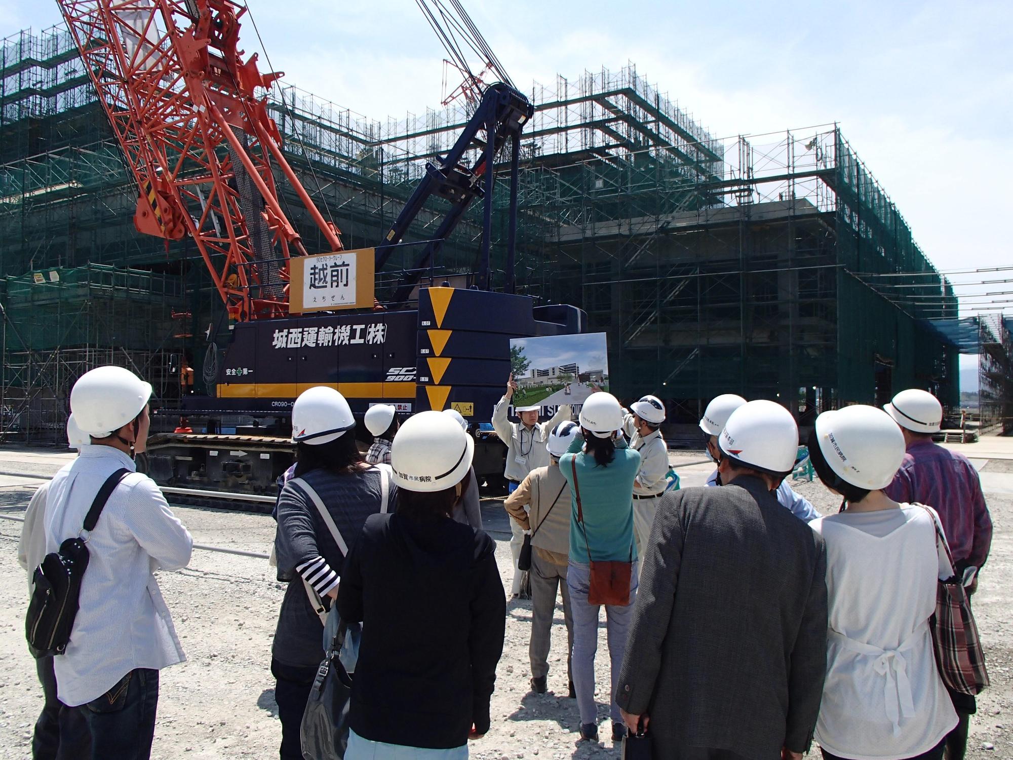 建設途中の建物の前で、資料を掲げて説明している作業服姿の人と、ヘルメットを着用して聞いている人たちの写真